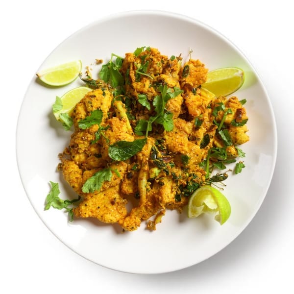 A white plate holds golden-brown spiced chicken pieces garnished with fresh cilantro and lime wedges.