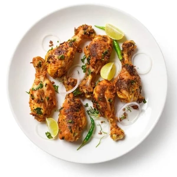 Six chicken drumsticks with herbs, lime wedges, green chilies, and onion rings on a white plate.