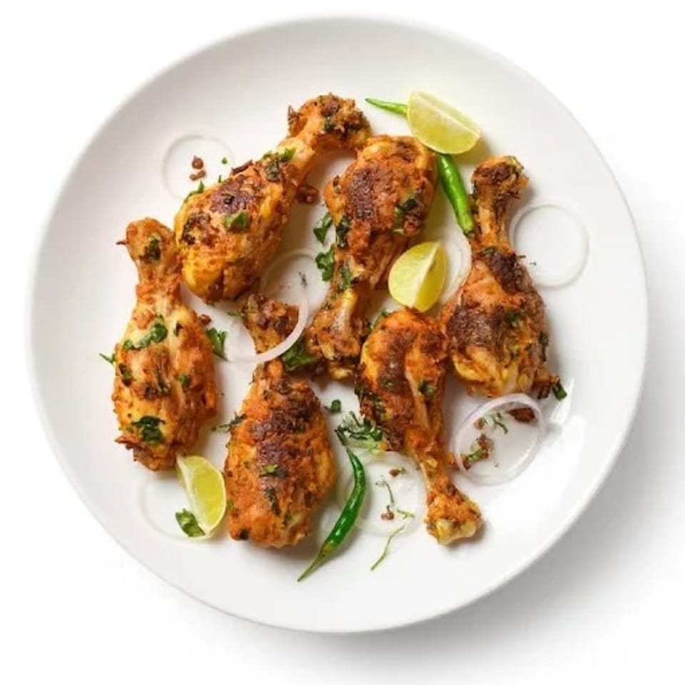Six chicken drumsticks with herbs, lime wedges, green chilies, and onion rings on a white plate.