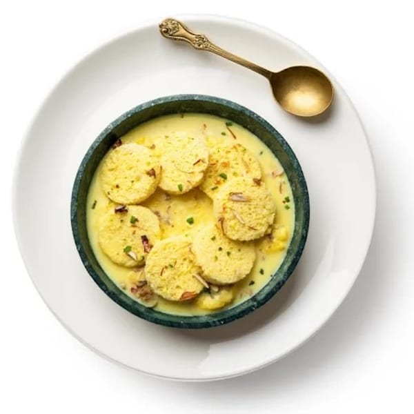 Rasmalai, an Indian dessert, in a dark bowl with a golden spoon on a white plate.